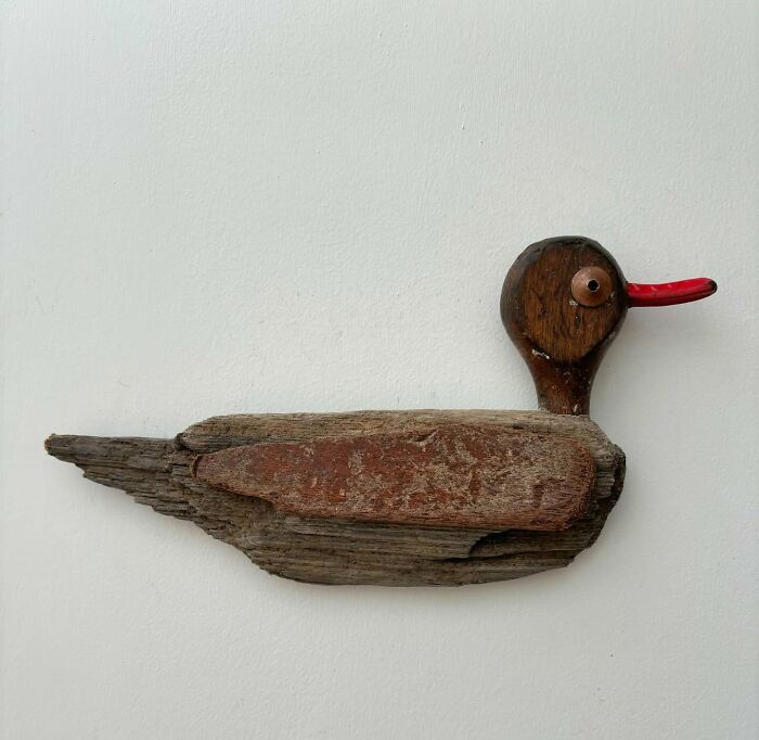 Driftwood animal sculpture resembling a duck with a red beak on a plain background.