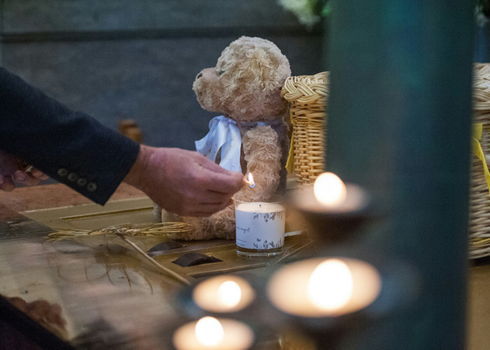 A person lights a candle next to a teddy bear in a somber setting, highlighting real-life insurance horror.