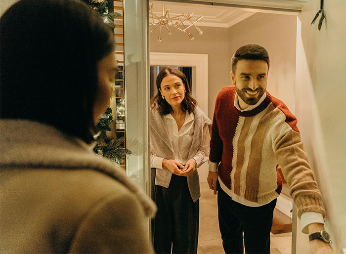 "Couple at door greeting guest, preparing for petty revenge in holiday setting.