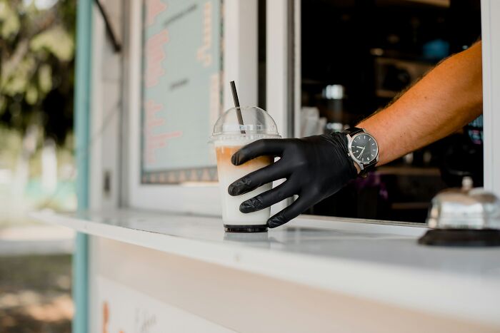 Barista with gloves serving iced coffee at a cafe window, reflecting modern hygiene practices.