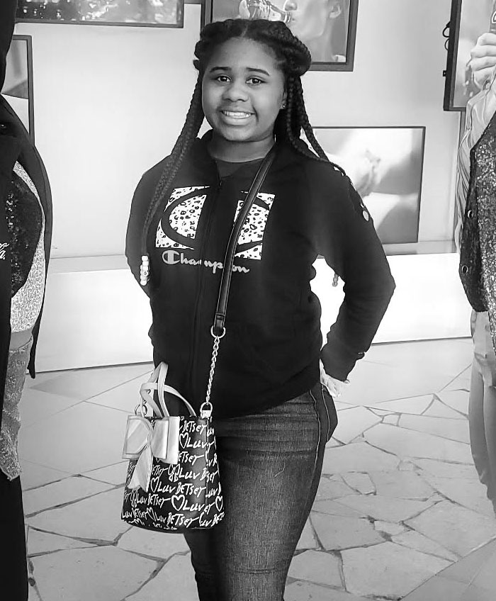 Kelaia Turner smiling in a casual setting, wearing a Champion hoodie and carrying a patterned bag, related to bullying and abuse. Kelaia Turner smiling in a casual setting, wearing a Champion hoodie and carrying a patterned bag, related to bullying and abuse.