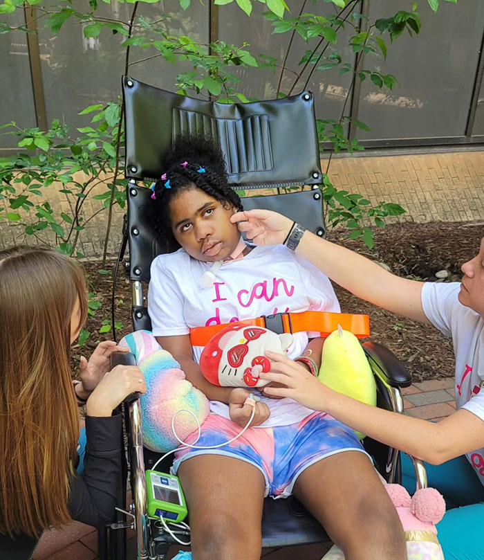 Kelaia Turner in a wheelchair outside, surrounded by supportive friends, holding plush toys. Kelaia Turner in a wheelchair outside, surrounded by supportive friends, holding plush toys.