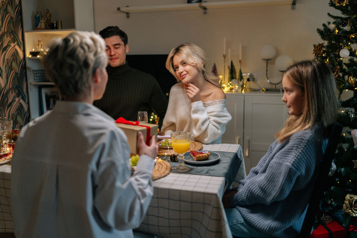 Family gathered at a festive table, exchanging gifts, highlighting kids' revenge themes amidst holiday decor.