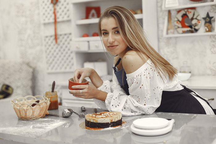 Young woman in a kitchen, holding a bowl, preparing dessert near a small cake.
