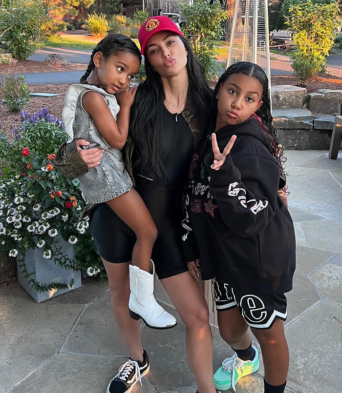 Kim Kardashian doing a silly pose in a red cap with two children, one being held, outside on a stone path near greenery with daughters Chicago and North West.
