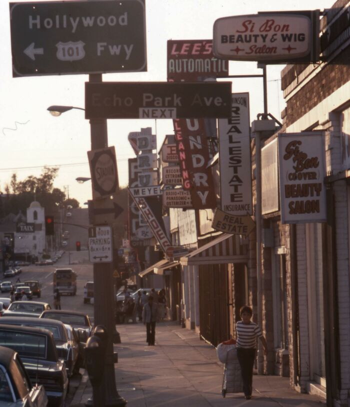 1970s West Coast street scene with vintage storefront signs and a person walking on the sidewalk under warm sunlight.