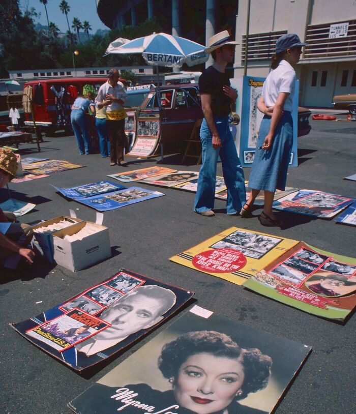 Vintage 1970s West Coast market scene with movie posters displayed on the ground, people browsing under a parasol.