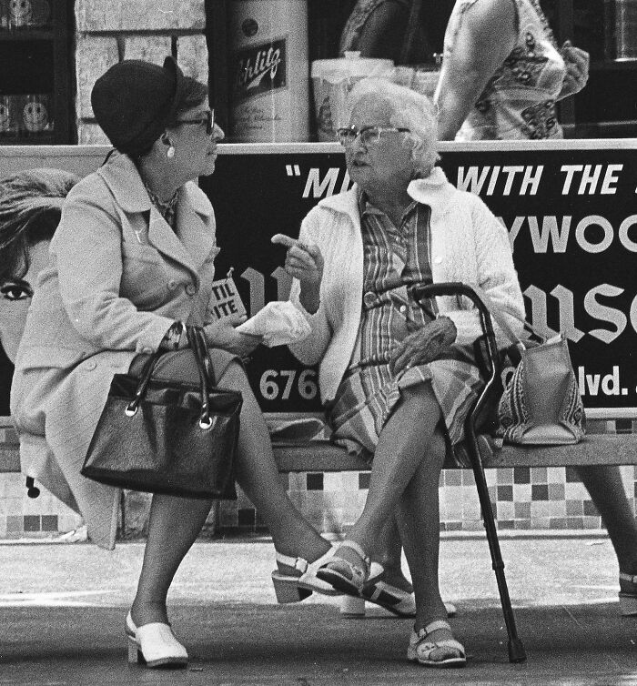 Two elderly women in 1970s West Coast fashion, chatting on a bench with vintage advertisements in the background.