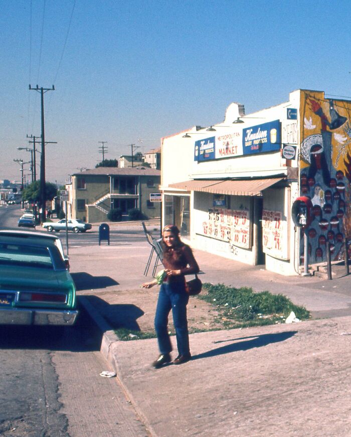 Person walking down a 1970s West Coast street, past a vintage market with colorful signage and classic cars.