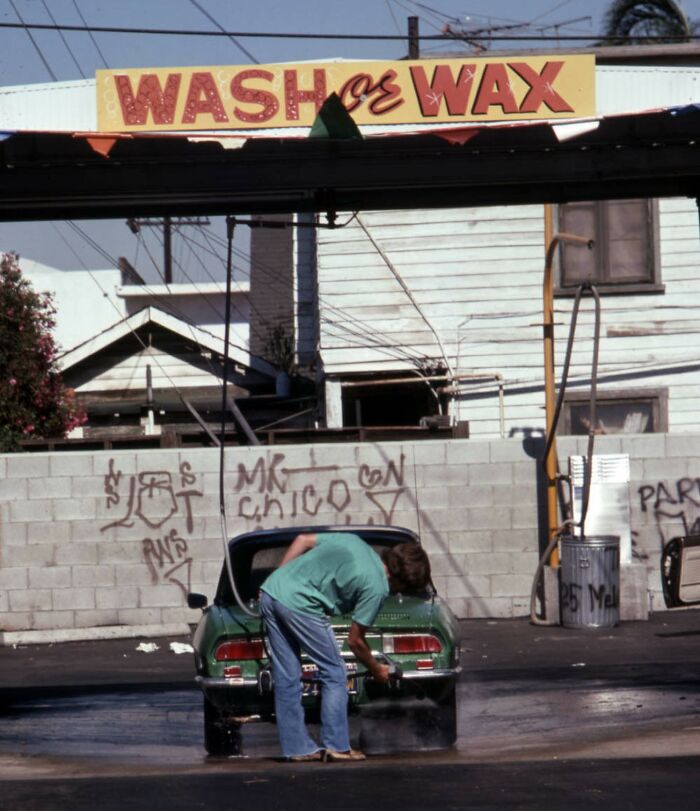 Man washing a vintage car at a 1970s West Coast car wash, with graffiti on the wall and a "Wash or Wax" sign above.