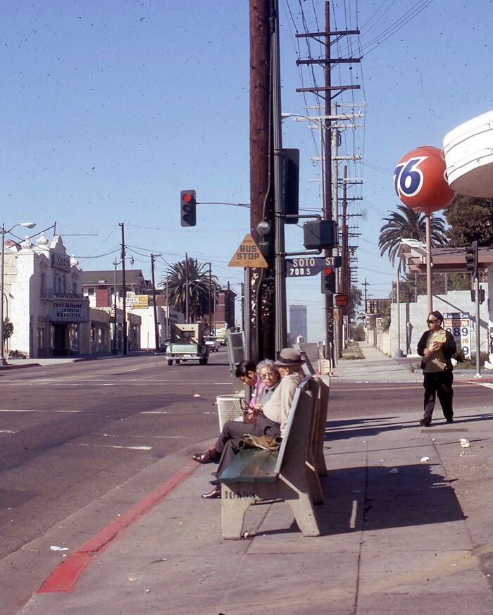 1970s West Coast street scene with people sitting at a bus stop, iconic old cars and a 76 gas station sign.