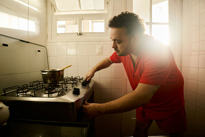 Man in red shirt adjusting stove knobs in a bright kitchen, sunlight streaming in.