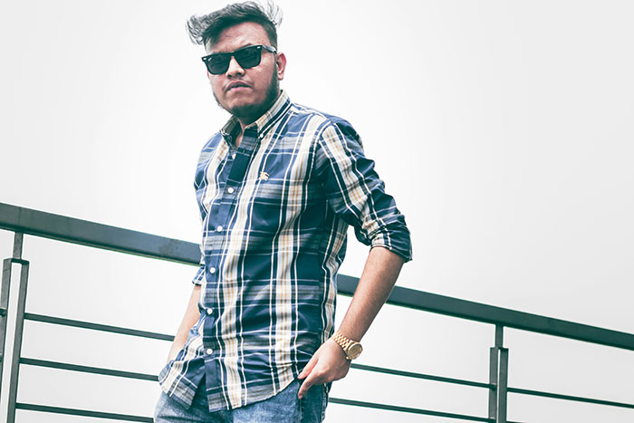 Man in sunglasses and plaid shirt posing casually by railing.