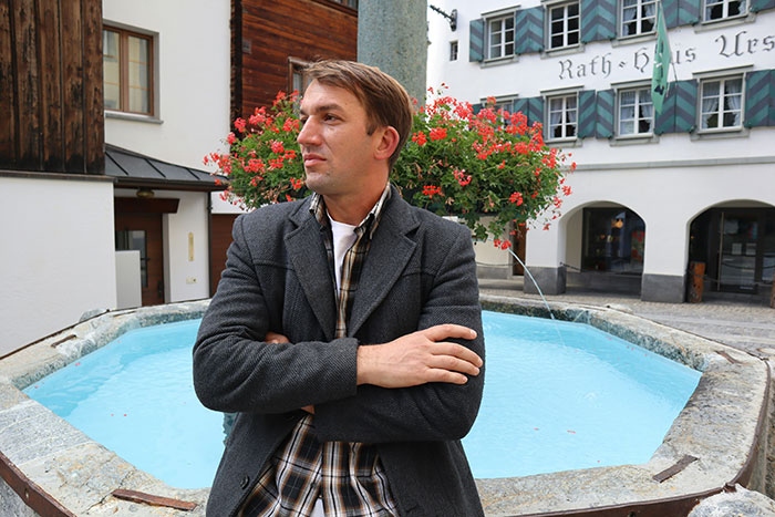 Man in a gray jacket standing with arms crossed by a decorative fountain, showcasing unattractive behavior in public spaces.