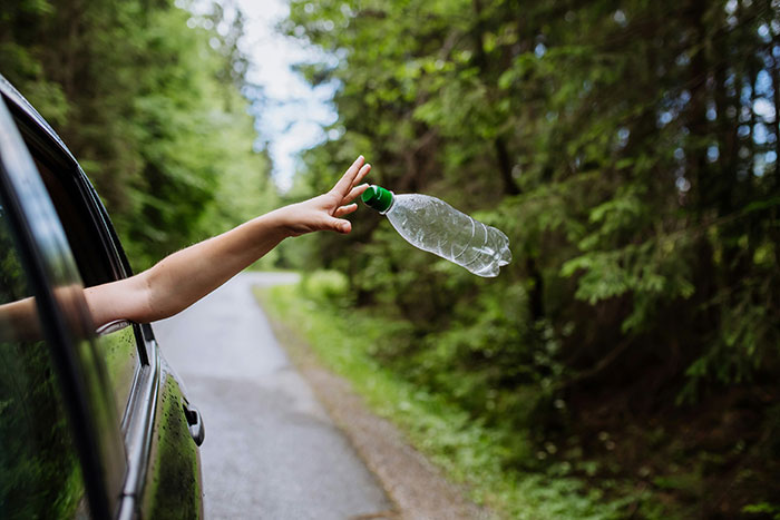 Person littering by throwing a plastic bottle from a car window into a forest area.