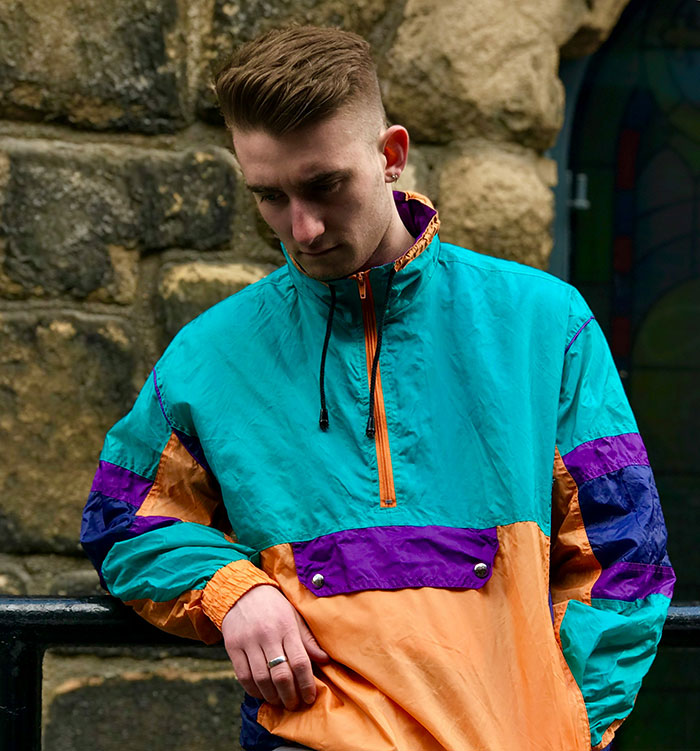 Person wearing a colorful windbreaker, standing against a stone wall, embodying unattractive fashion choices.