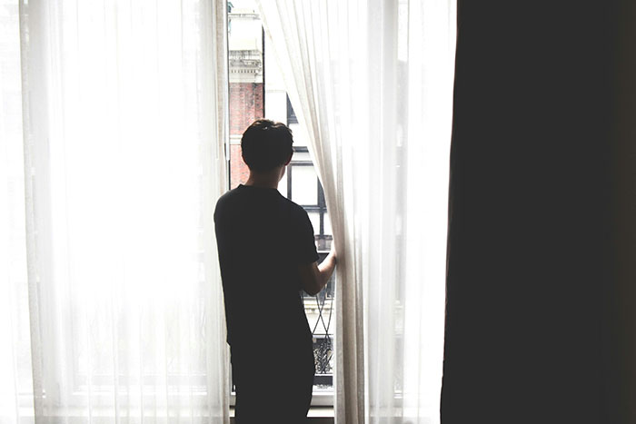 Person standing alone by a window, partially obscured by curtains, illustrating unattractive habits.