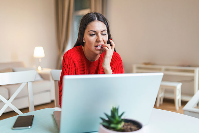 Woman in red sweater biting nails, an unattractive habit, while using a laptop in a cozy living room.