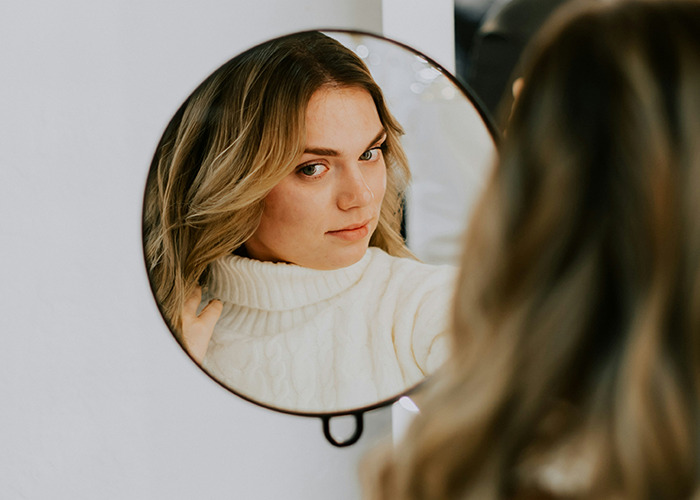 Woman in a cozy white sweater looking at herself in a round mirror, reflecting on health hacks.