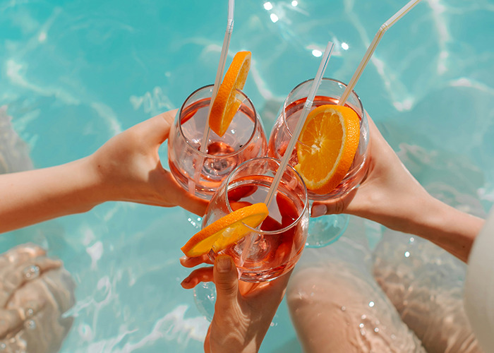 Three hands holding cocktail glasses with orange slices over a swimming pool, representing health hacks and relaxation.