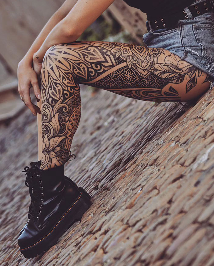 A detailed mandala tattoo design on a person's leg, wearing shorts and boots, leaning on a brick surface.