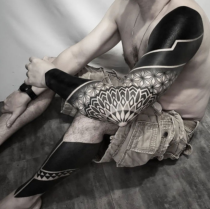 Man with intricate mandala tattoos on arm and leg, showcasing geometric designs.