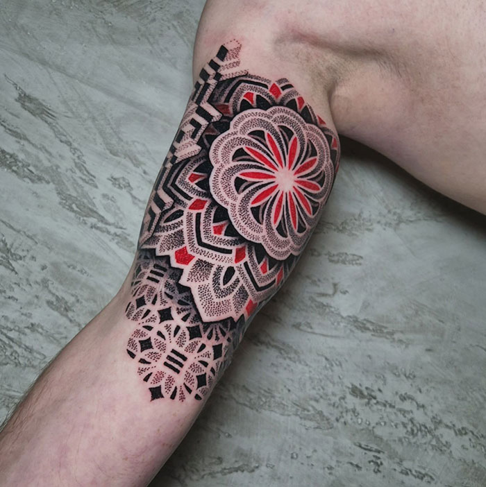 Intricate mandala tattoo in red and black ink on upper arm, showcasing detailed geometric patterns.
