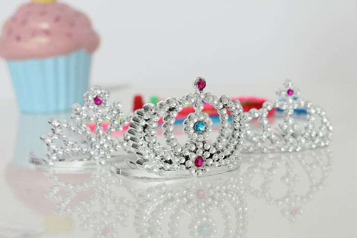 Three silver tiaras with colorful gemstones on a reflective surface, cupcake decoration in the background.