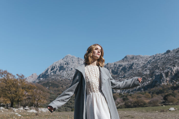 Woman in a scenic mountain setting, wearing a white dress and gray coat, embodying Pinterest trends for 2025.