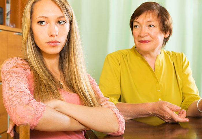 New mom looking upset, arms crossed, sitting next to an older woman in a yellow shirt, inside a home environment. New mom looking upset, arms crossed, sitting next to an older woman in a yellow shirt, inside a home environment.