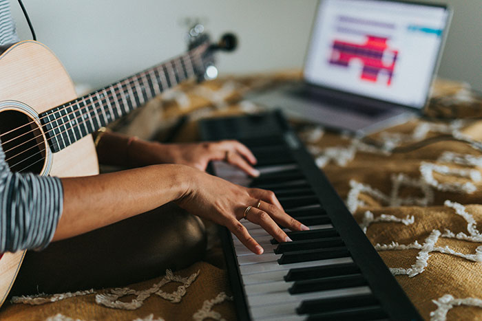 Person playing keyboard with a guitar nearby, highlighting the gifted stereotype. Laptop in the background on a patterned bedspread.