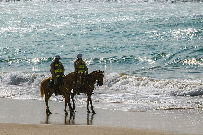 Mounted police patrol a beach, with waves crashing nearby, under a clear sky.