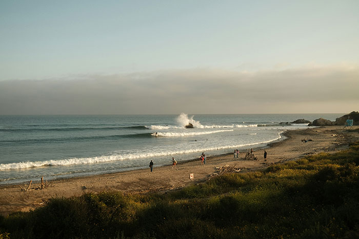 People walking along a beach with waves crashing on rocks in the distance.