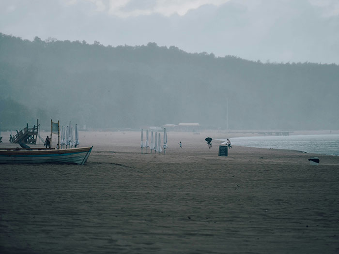 Foggy beach scene with a few people walking, umbrellas closed, and a boat on the sand.
