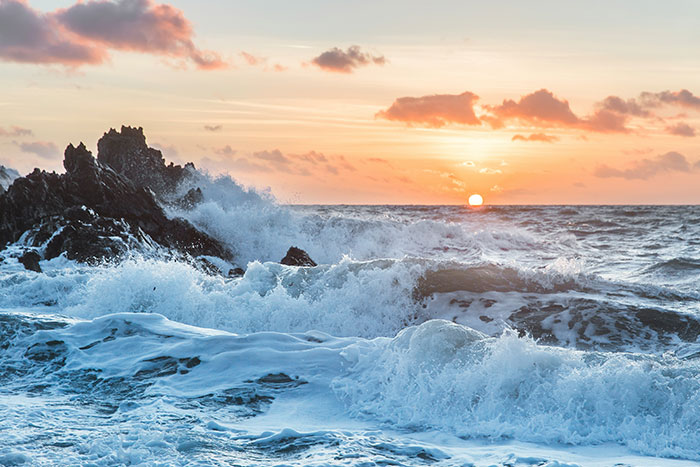 Sunset over crashing waves at the beach, with dramatic clouds and rocky outcrops.