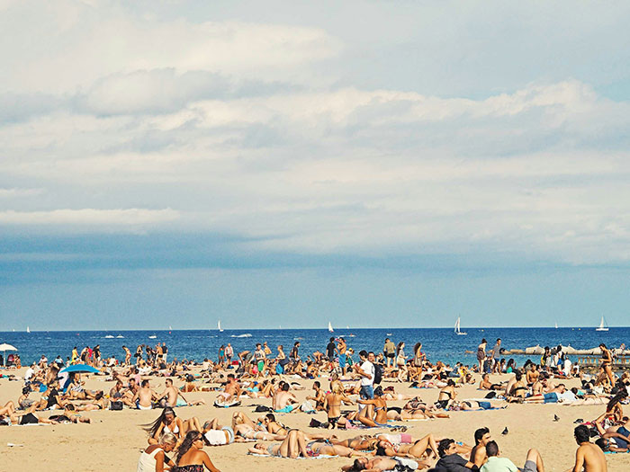 Crowded beach scene with people sunbathing and sailing in the background, capturing a typical summer day at the seaside.