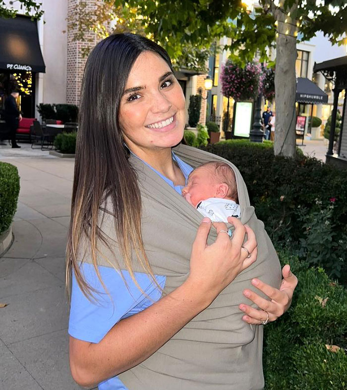 Momfluencer smiling outdoors, holding a sleeping newborn wrapped in a sling.