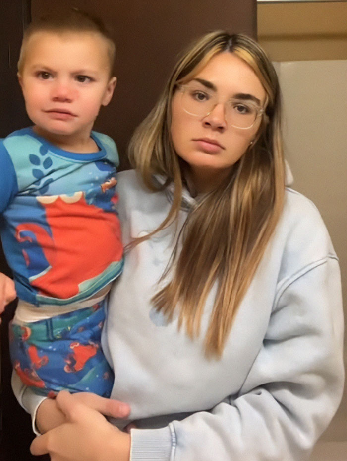 Momfluencer holding child with a serious expression, wearing glasses and a hoodie, amid controversy and investigation.