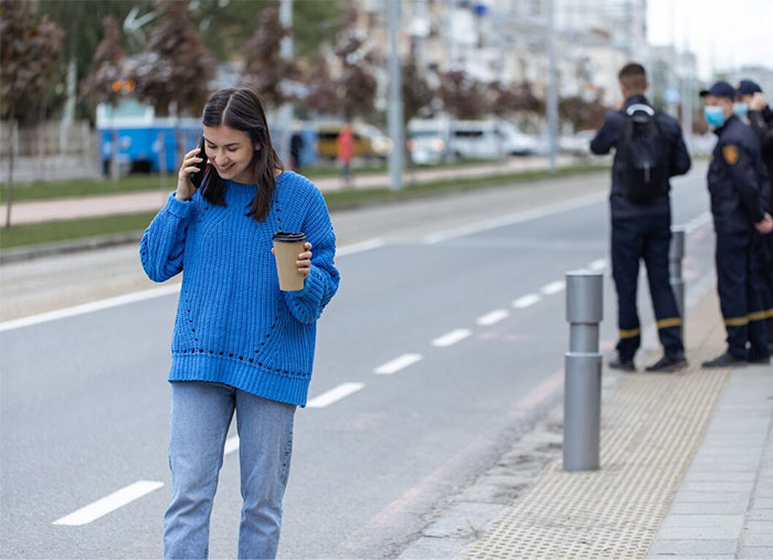 Woman in a blue sweater talking on the phone, holding coffee, on a street.