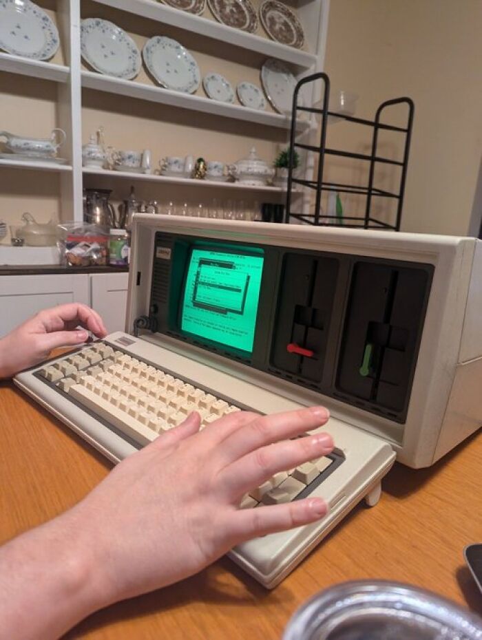 Hands typing on a vintage portable computer, evoking an old internet feel with a green text screen display.
