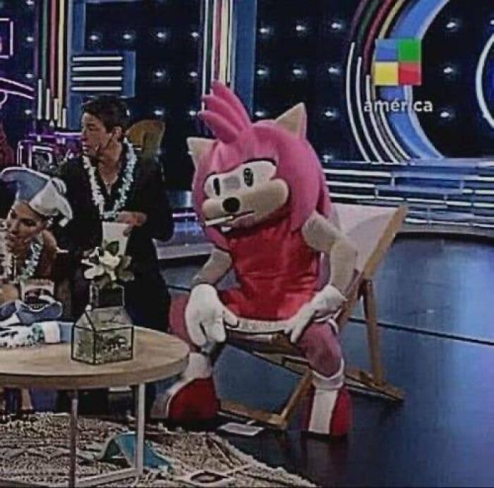 Person in a pink cartoon costume sitting on a TV set, evoking old internet feel vibes.