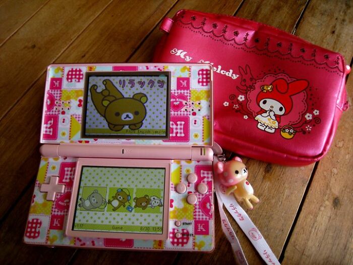 Old-Internet-Feel-Pics: A colorful Nintendo DS featuring Rilakkuma next to a My Melody pouch on a wooden table.