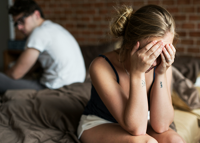 Woman upset on bed; fianc&eacute; turns away after touchy behavior dispute with female friend.