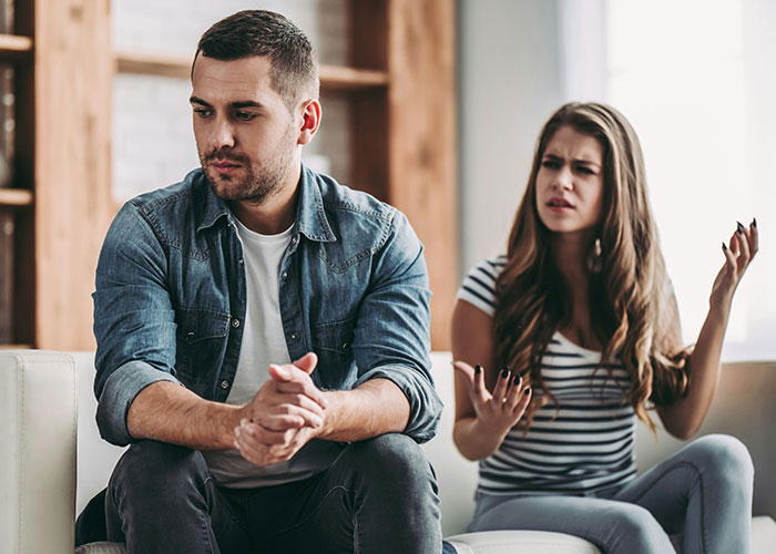 Woman confronts fianc&eacute; about behavior with friend, expressing frustration while he looks away, sitting in a living room.