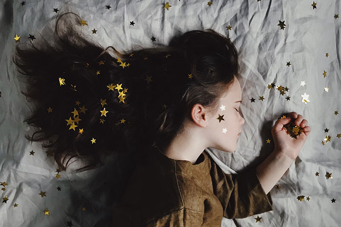 Child lying on a bed with hair adorned by golden stars, reflecting a dreamy Hollywood secrets theme.