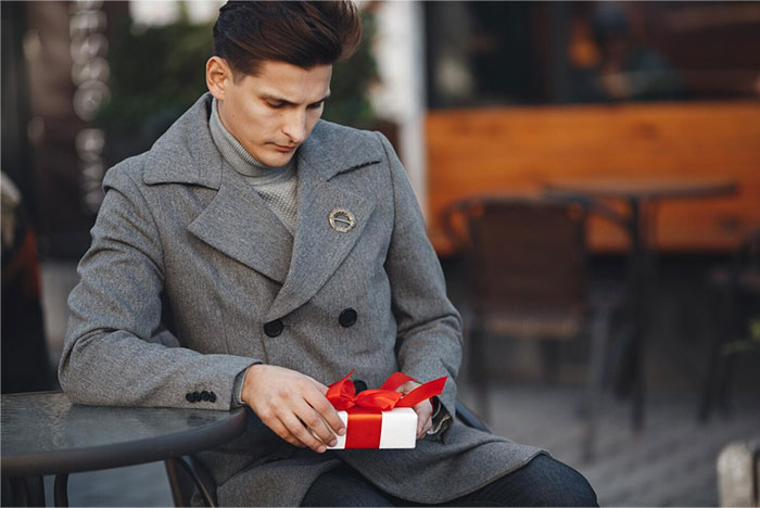 Man in gray coat sitting at a table, holding a small gift box with a red ribbon, contemplating Hollywood secrets.