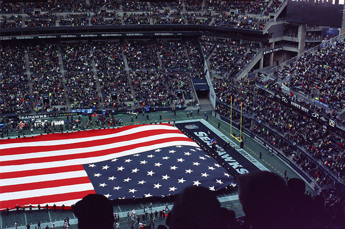 Large American flag displayed on a football field during a packed event, surrounded by thousands of spectators.
