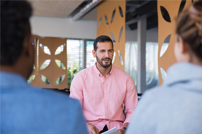 Man in a pink shirt engaging in discussion, sharing Hollywood secrets with two people in a modern office setting.