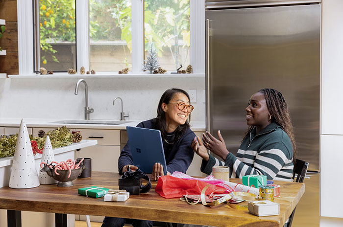 Two people at a kitchen table, smiling and talking, possibly discussing Hollywood secrets amidst gift wrapping supplies.
