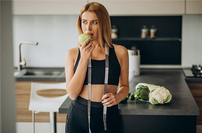 Woman in a kitchen holding an apple, wearing a sports bra and measuring tape, suggesting Hollywood secrets.